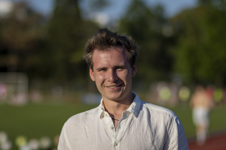 Junger Mann mit einem lächelnden Gesicht, in weißem Hemd, steht im Freien auf einem Sportplatz im Hintergrund.