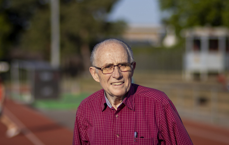 Älterer Mann mit Brille, der auf einem Sportplatz steht, lächelt in die Kamera. Im Hintergrund sind Sportanlagen sichtbar.