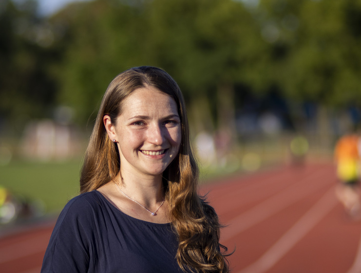 Lächelnde Frau mit langen, braunen Haaren steht auf einer Laufbahn, unscharfer Hintergrund mit Sportlern.