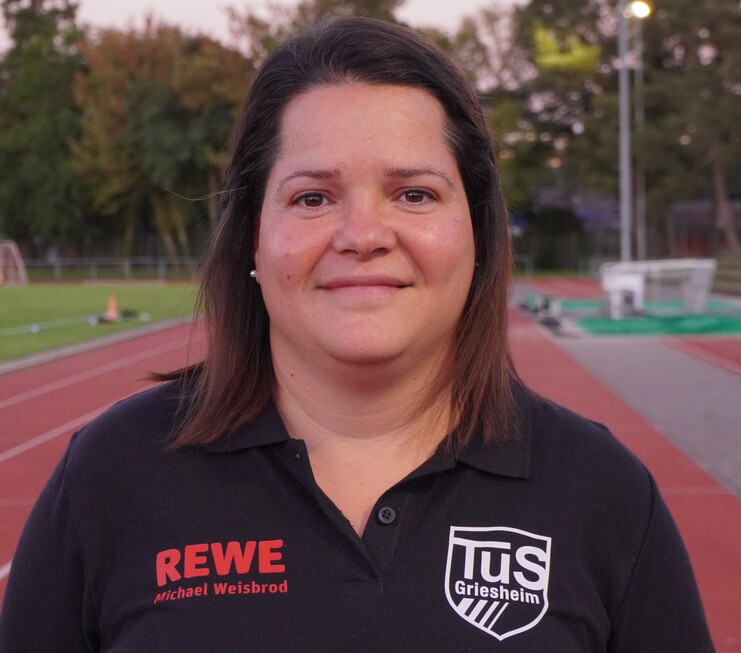 Porträt einer Frau in einem schwarzen Polo-Shirt mit Vereinslogo von TuS Griesheim, Hintergrund mit Sportplatz.