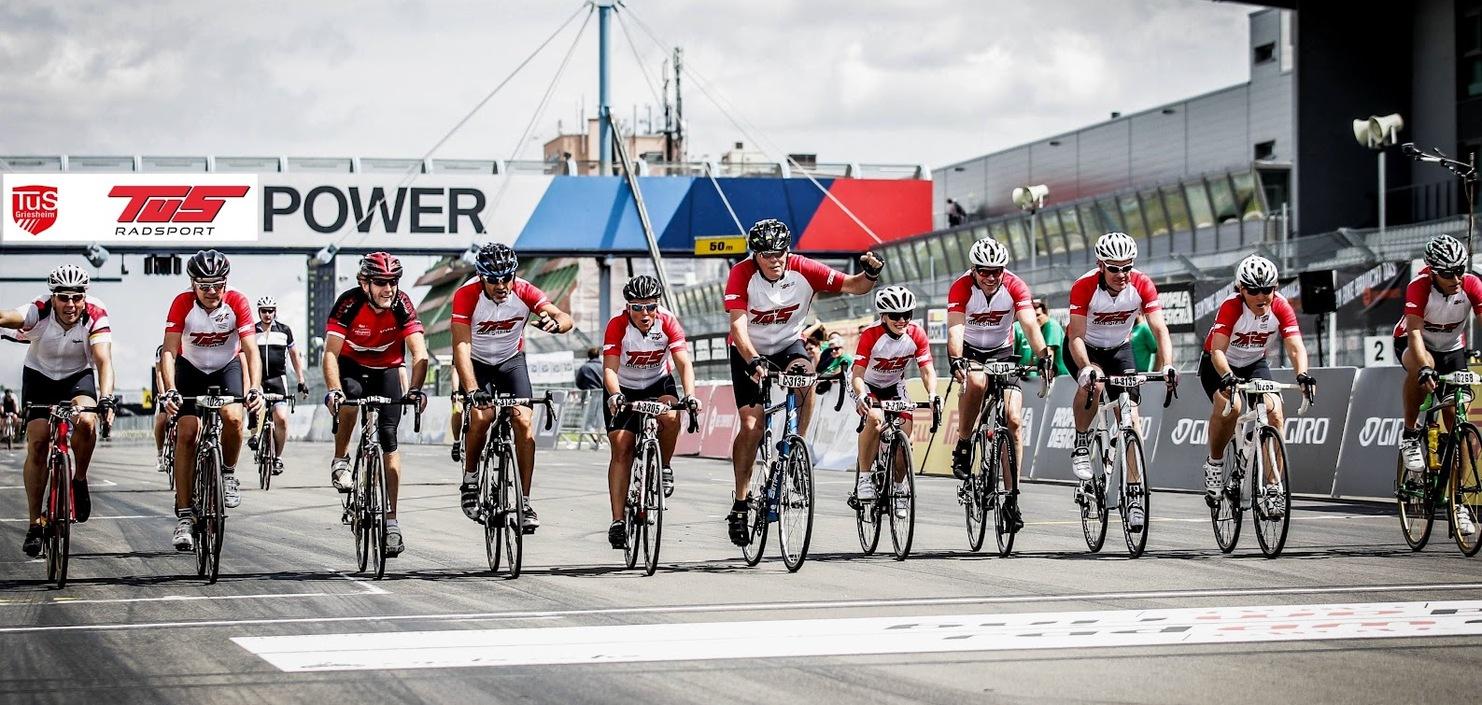Gruppierung von Radfahrern in roten Shirts, die die Ziellinie auf einer Rennstrecke &uuml;berqueren.