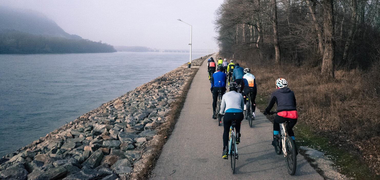 Gruppe von Radfahrern auf einem Weg entlang eines Flusses, umgeben von Bäumen und Nebel.