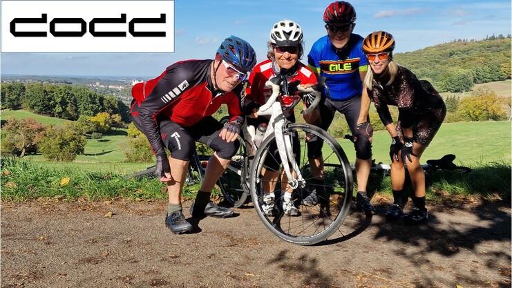 Gruppe von vier Radfahrern posiert mit einem Rennrad und einem platten Reifen vor einer grünen Landschaft.