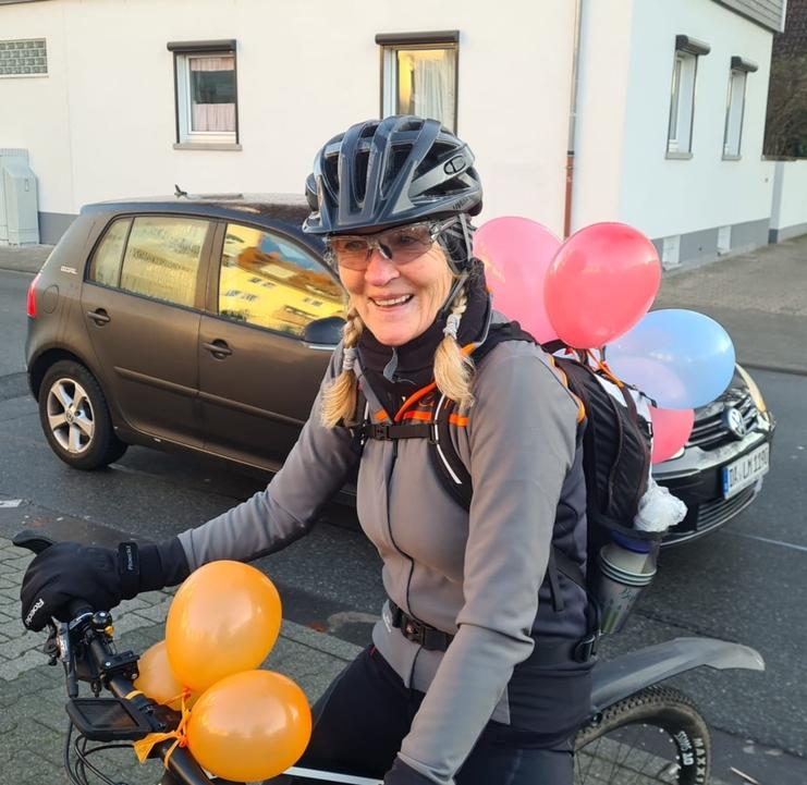 Frau mit Zöpfen und Fahrradhelm auf einem Fahrrad, umgeben von bunten Luftballons, lächelt in die Kamera.