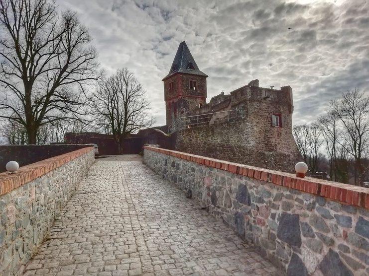 Historische Burg mit Turm, umgeben von Bäumen, auf einem gepflasterten Weg mit Mauer und laternenartiger Beleuchtung.