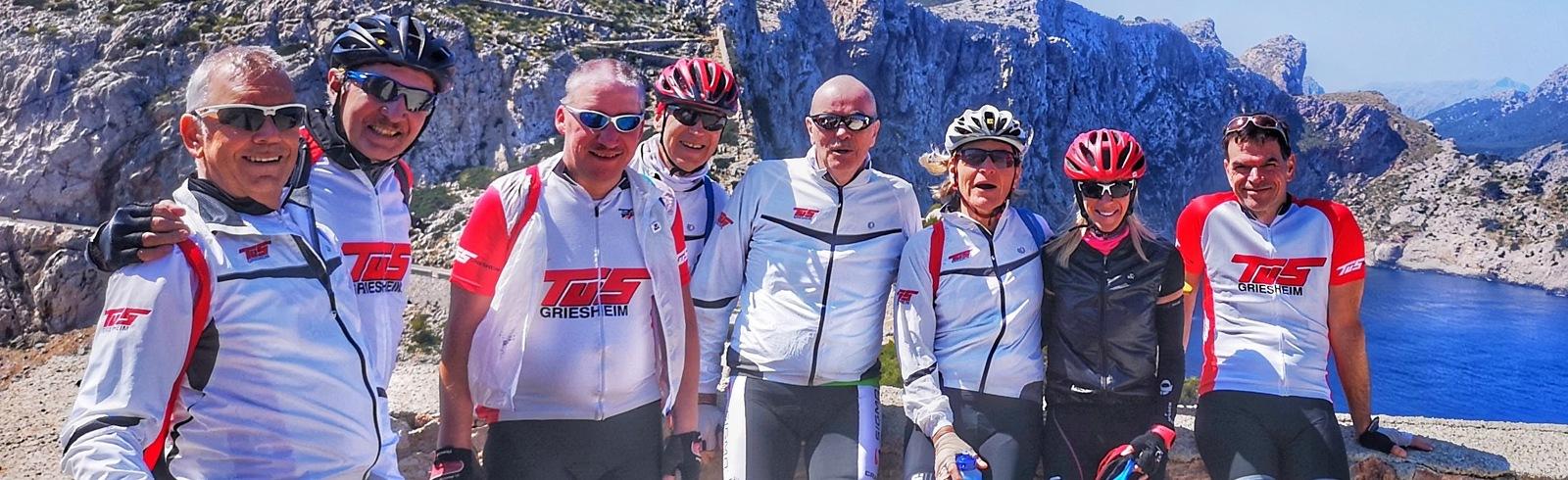 Gruppe von neun Radfahrern in Sportkleidung posiert vor einer K&uuml;stenlandschaft mit Bergen und blauem Wasser.