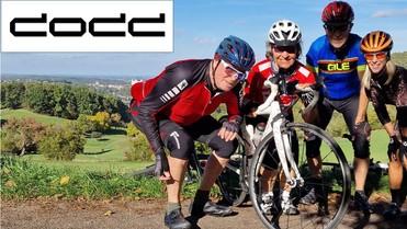 Gruppe von vier Radfahrern posiert mit einem Rennrad und einem platten Reifen vor einer grünen Landschaft.