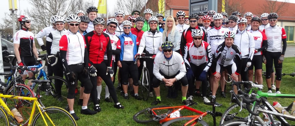 Gruppennahaufnahme von Radfahrern in Sportkleidung mit Fahrrädern auf einer Wiese vor einem Verbrauchermarkt.