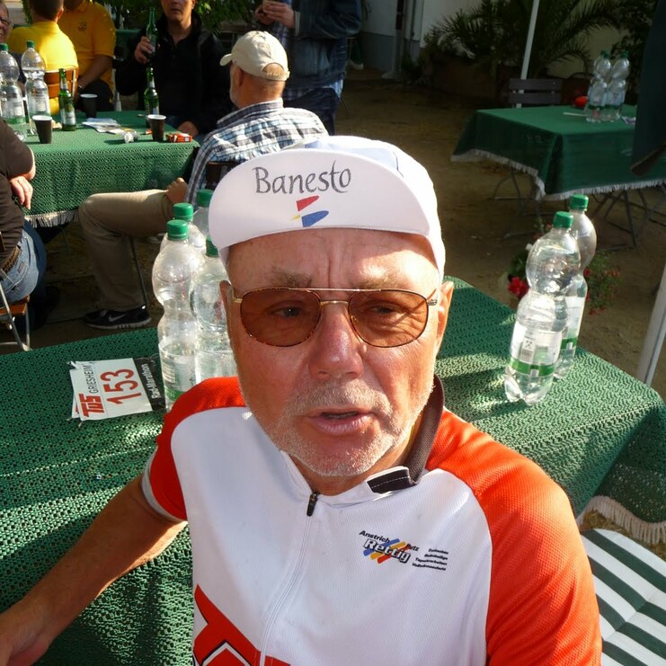 Älterer Radfahrer mit Sonnenbrille und Kappe sitzt an einem Tisch, umgeben von anderen Personen und Flaschen.