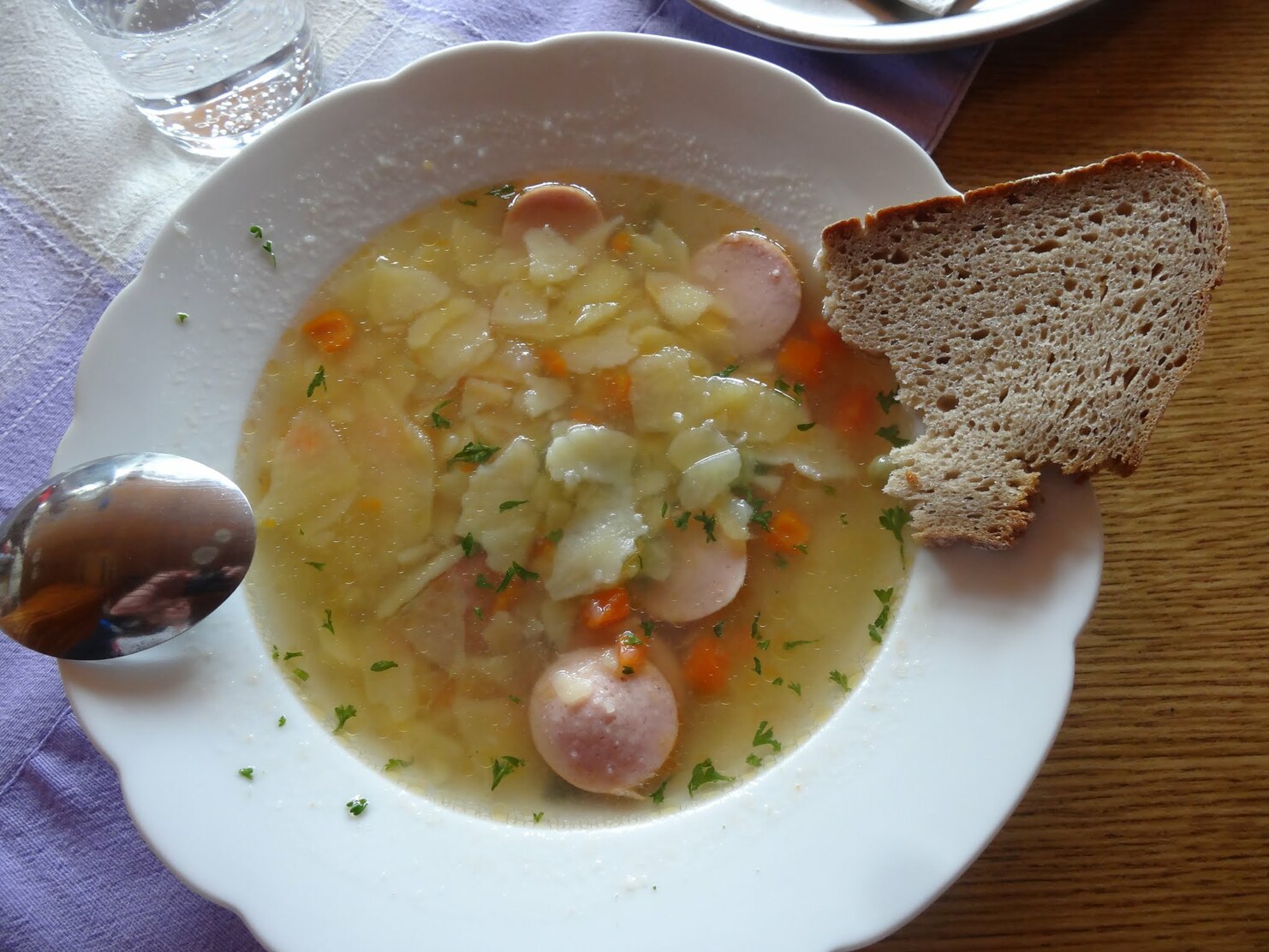 Schüssel mit Suppe aus Gemüse und Würstchen, daneben ein Stück Roggenbrot auf einem Holztisch.