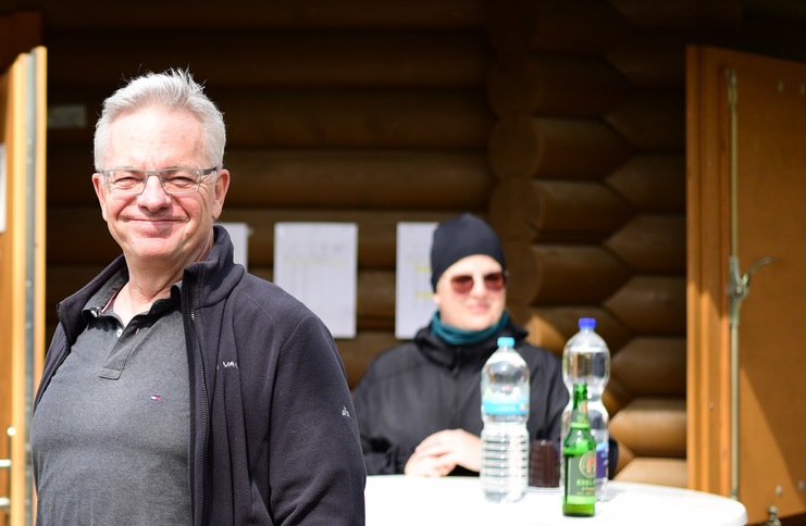 L&auml;chelnder Mann im Vordergrund, im Hintergrund eine Person mit Sonnenbrille und Getr&auml;nken an einem Tisch.