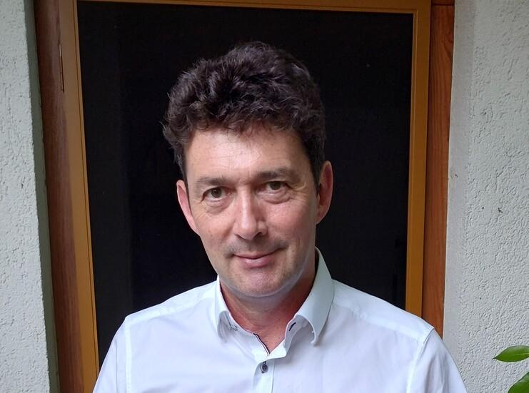 Porträt eines Mannes mit dunklen Locken, der in einem Raum vor einem Fenster steht. Er trägt ein helles Hemd.