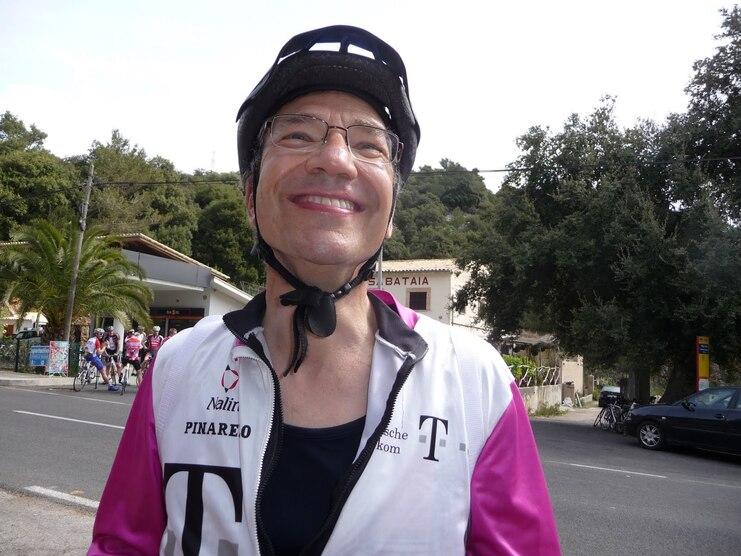 Lächelnde Person in Radfahrerkleidung und Helm vor einer Straße mit Bäumen und Gebäuden im Hintergrund.