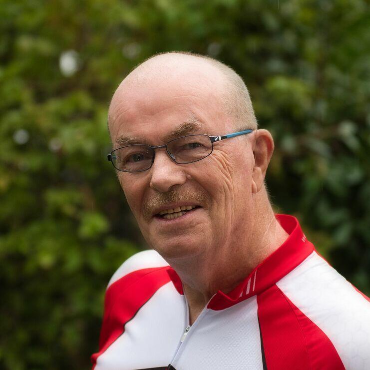 Lächelnder Mann in Brille trägt ein rot-weißes Fahrradtrikot vor grünem Hintergrund.