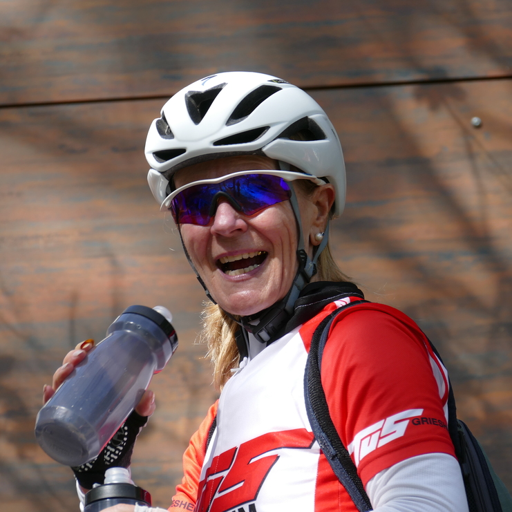 Frau in Radrennausrüstung lächelt, hält eine Wasserflasche und trägt einen Fahrradhelm mit Sonnenbrille.