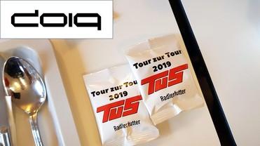 Zwei Tütchen mit dem Aufdruck „Tour zur Tour 2019“ und „Radlerfütter“ liegen neben Besteck auf einem Tisch.