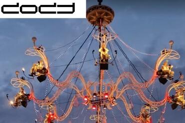 Großer, leuchtender Kronleuchter mit Künstlern, beleuchtet von bunten Lichtern, vor einem bewölkten Abendhimmel.