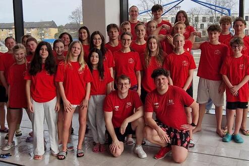 Gruppe von Jugendlichen und Betreuern in roten T-Shirts, posierend vor einem Fenster in einem Schwimmbad.