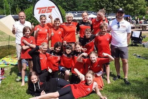 Gruppe von Kindern und Betreuern in roten T-Shirts, lächelnd im Freien vor Zelt und Bäumen, einige zeigen Daumen hoch.