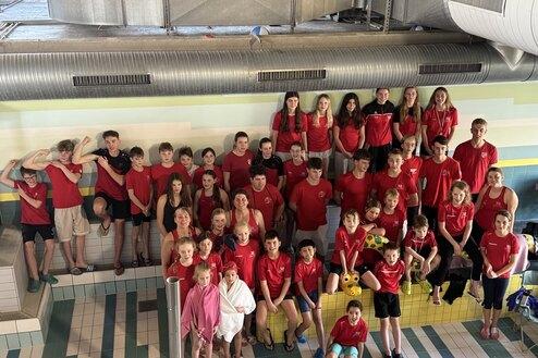 Gruppenshot von 44 Schwimmern in roten Shirts, lächelnd vor einer bunten Wand im Schwimmbad.