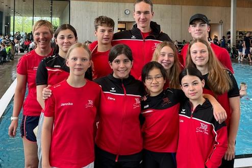 Gruppe junger Schwimmer in roten Trainingsanz&uuml;gen, l&auml;chelnd vor einem Schwimmbecken und Holzstruktur im Hintergrund.