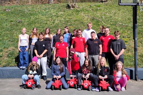 Gruppe von Jugendlichen posiert im Freien mit roten Spielautos vor einem Basketballkorb und einer grünen Wiese.