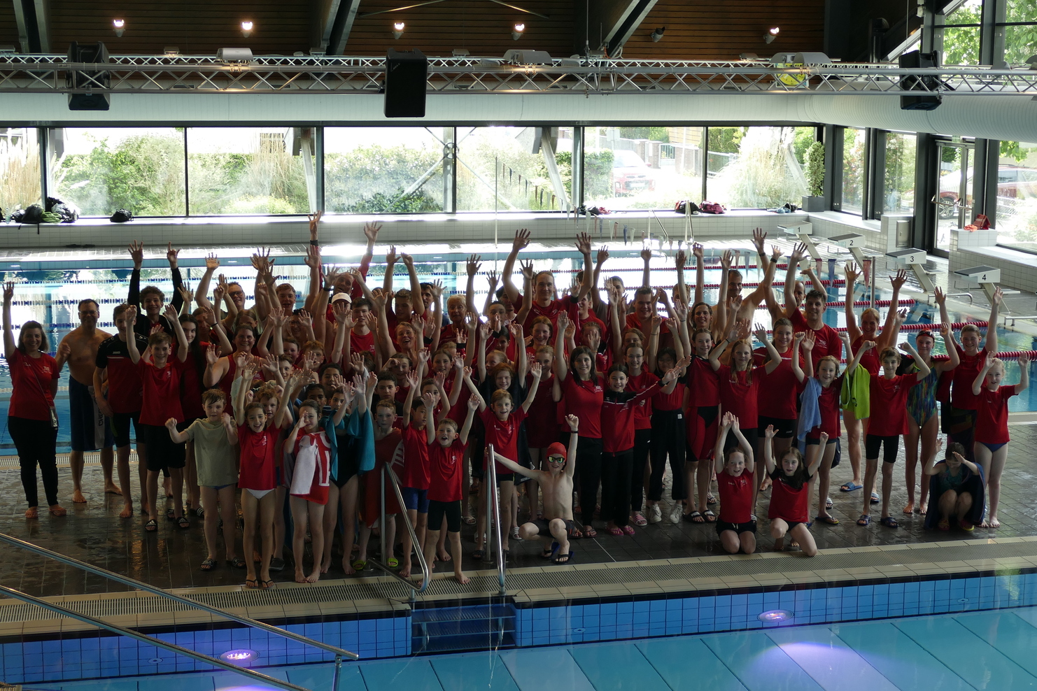Gruppenfoto von Schwimmern in roten T-Shirts, die jubelnd am Beckenrand eines Schwimmbads stehen.