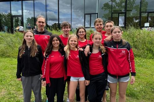 Gruppe von zehn Jugendlichen in roten Sportanz&uuml;gen, stehend auf einer Wiese vor einem modernen Geb&auml;ude.