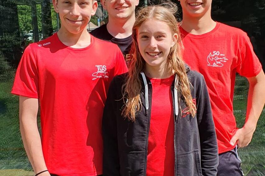 Vier Jugendliche in roten T-Shirts stehen vor einem großen Fenster und lächeln in die Kamera. Sonnenlicht und grüne Bäume im Hintergrund.