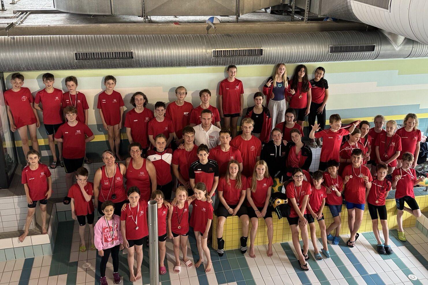 Gruppe von Schwimmern in roten T-Shirts posiert lächelnd am Beckenrand in einem Hallenbad.