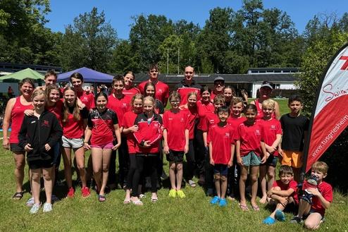 Gruppe von Schwimmern in roten T-Shirts steht auf einer Wiese, im Hintergrund B&auml;ume und Zelte unter blauem Himmel.