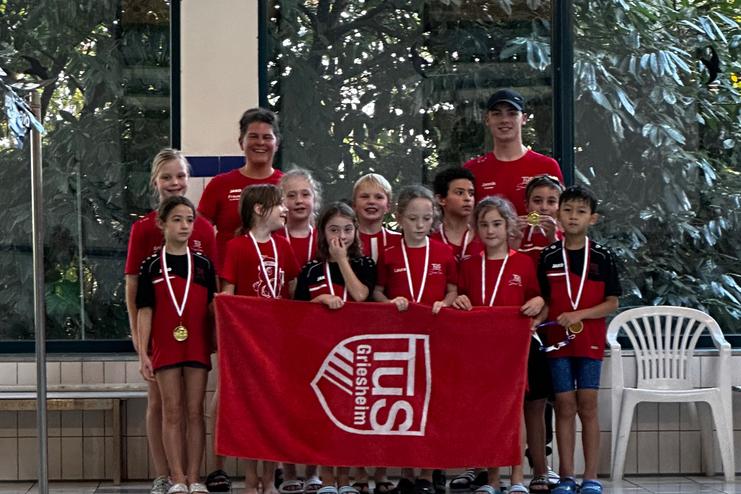 Gruppe junger Schwimmer in roten T-Shirts mit Medaillen, posiert vor einer Schwimmhalle mit einer Fahne.