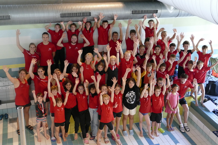 Gruppennfoto von Schwimmern in roten T-Shirts, die mit erhobenen Händen vor einem Schwimmbad posieren.