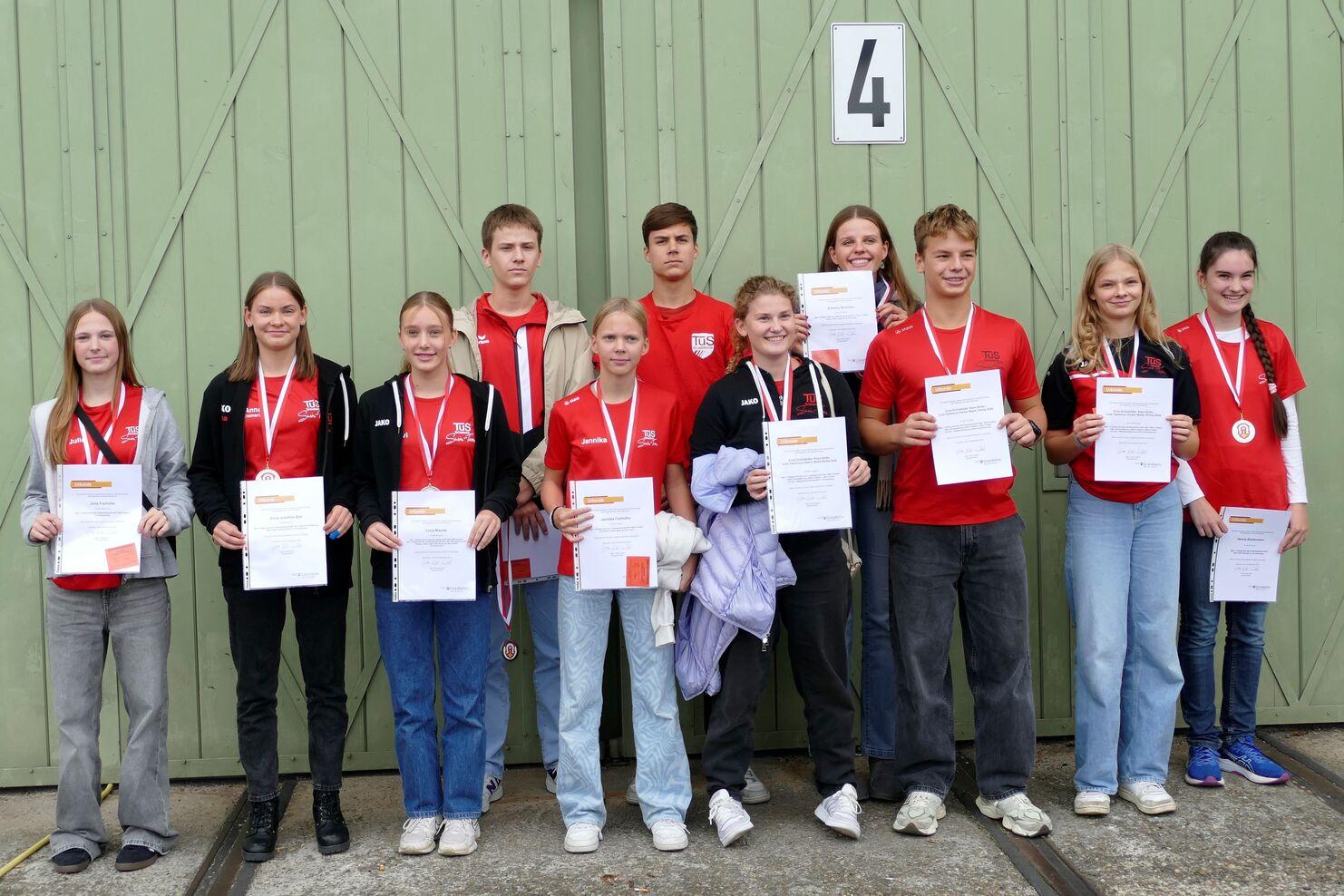 Gruppe von zehn Jugendlichen in roten T-Shirts mit Urkunden und Medaillen vor grüner Wand, lächelnd und posierend.