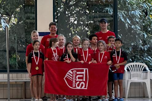 Gruppe junger Schwimmer in roten T-Shirts mit Medaillen, posiert vor einer Schwimmhalle mit einer Fahne.
