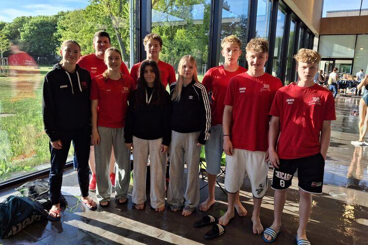 Gruppe von neun Schwimmern in roten T-Shirts, stehend im Freien vor großen Glasfenstern.