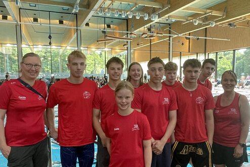 Gruppe von Schwimmern und Trainern in roten T-Shirts, stehend am Rand eines Hallenbades mit Schwimmbecken im Hintergrund.