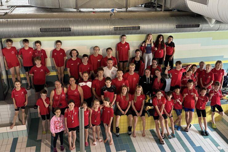 Gruppe von Schwimmern in roten T-Shirts posiert lächelnd am Beckenrand in einem Hallenbad.