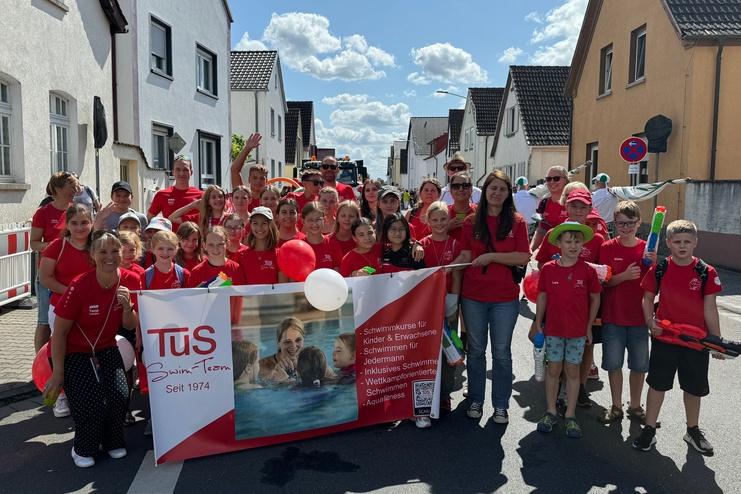 Gruppe von Kindern und Erwachsenen in roten T-Shirts mit Banner bei einem Umzug auf einer sonnigen Straße.