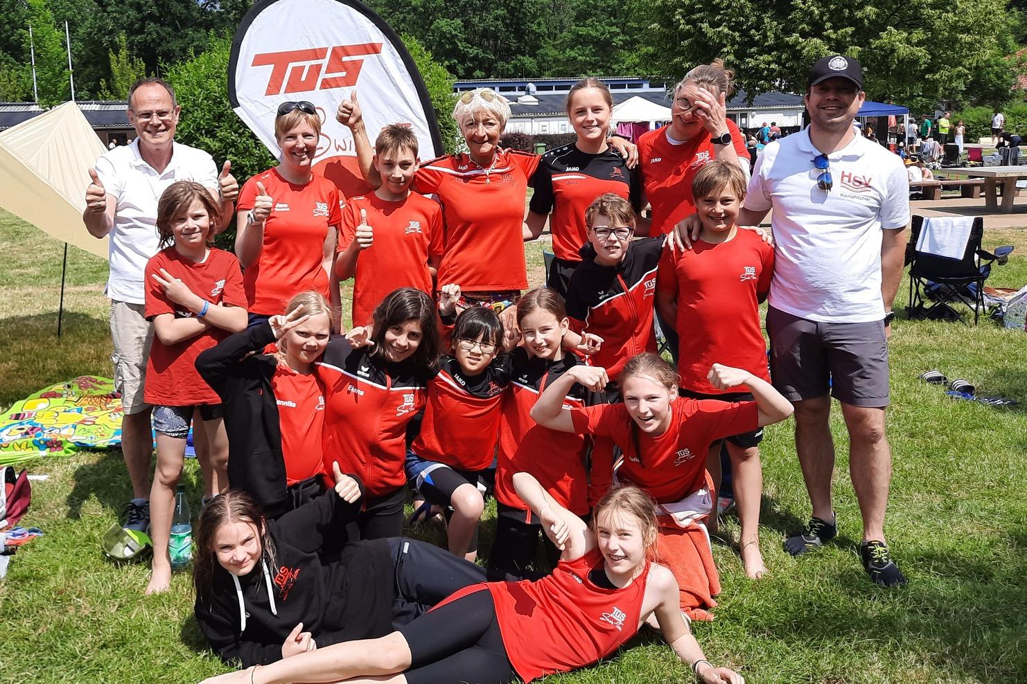 Gruppe von Kindern und Betreuern in roten T-Shirts, lächelnd im Freien vor Zelt und Bäumen, einige zeigen Daumen hoch.