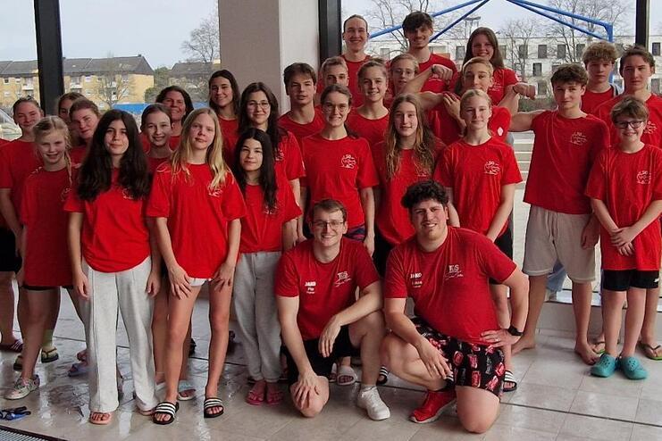 Gruppe von Jugendlichen und Betreuern in roten T-Shirts, posierend vor einem Fenster in einem Schwimmbad.