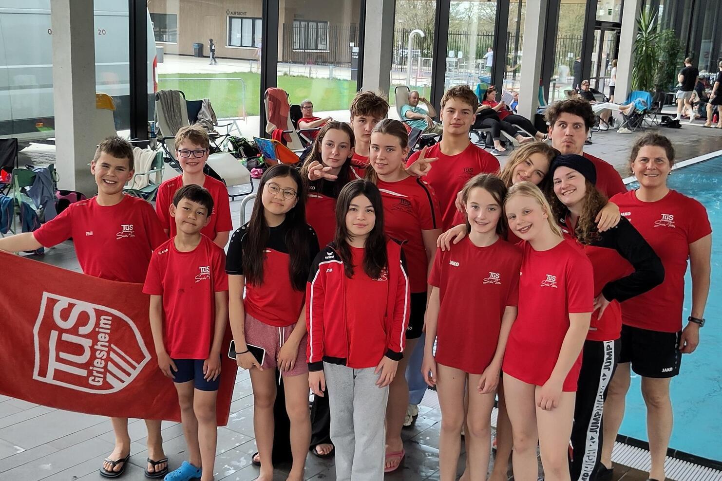 Gruppe junger Schwimmer in roten T-Shirts mit Vereinslogo TUS Griesheim, lächeln am Schwimmbeckenrand.