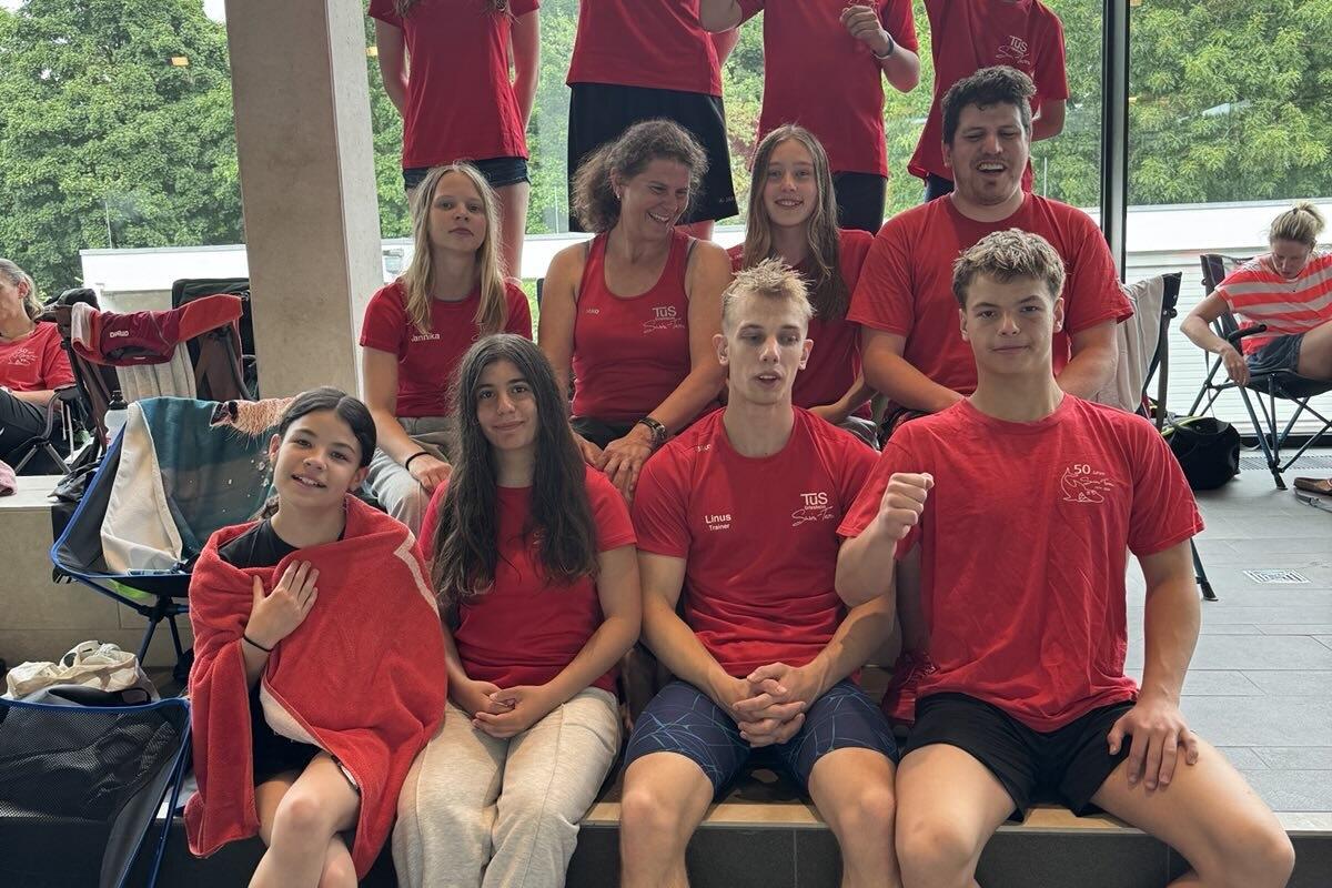 Gruppe von Schwimmern in roten T-Shirts posiert fröhlich vor einem Fenster im Schwimmbad.