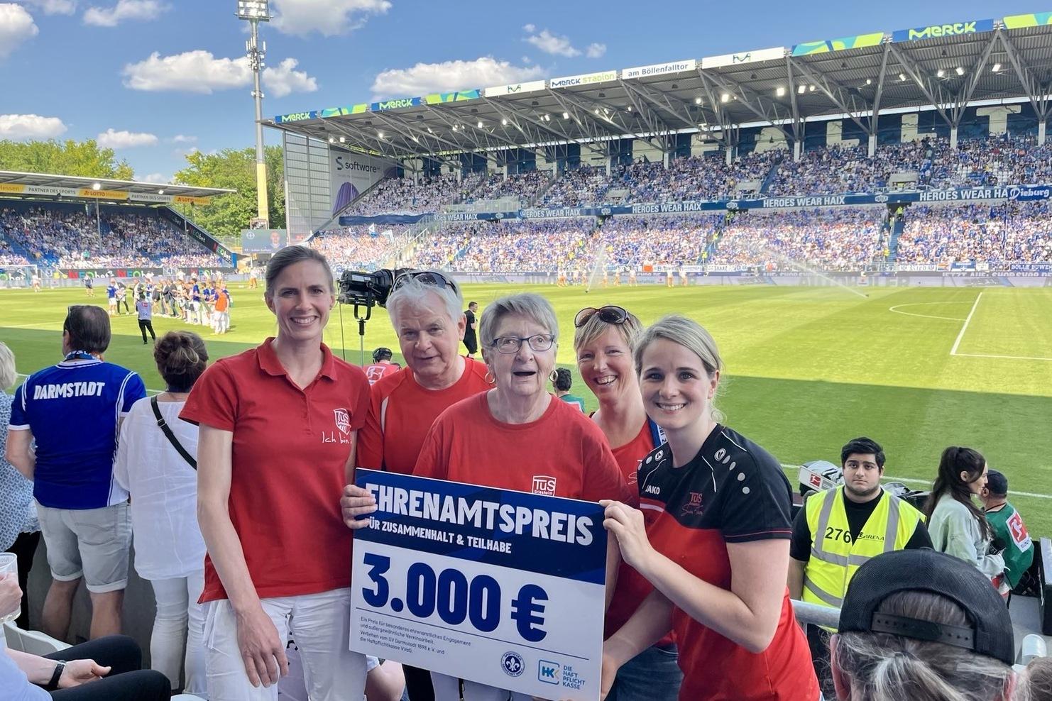 Vier Personen halten ein Schild mit der Aufschrift „Ehrenamtspreis 3.000 €“ in einem Fußballstadion. Hintergrund: Spielszene.