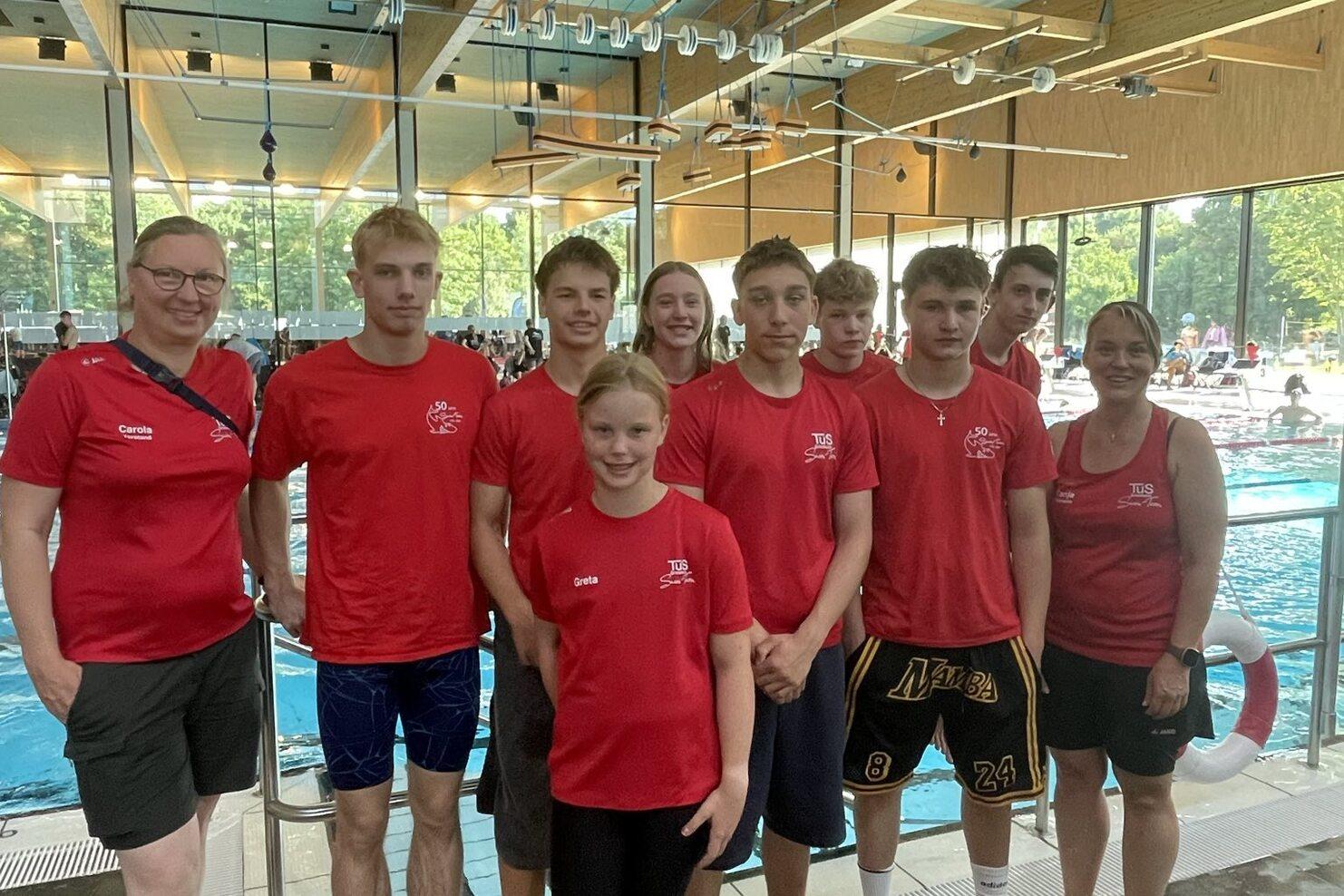 Gruppe von Schwimmern und Trainern in roten T-Shirts, stehend am Rand eines Hallenbades mit Schwimmbecken im Hintergrund.