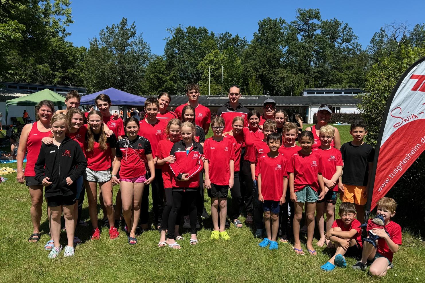 Gruppe von Schwimmern in roten T-Shirts steht auf einer Wiese, im Hintergrund Bäume und Zelte unter blauem Himmel.