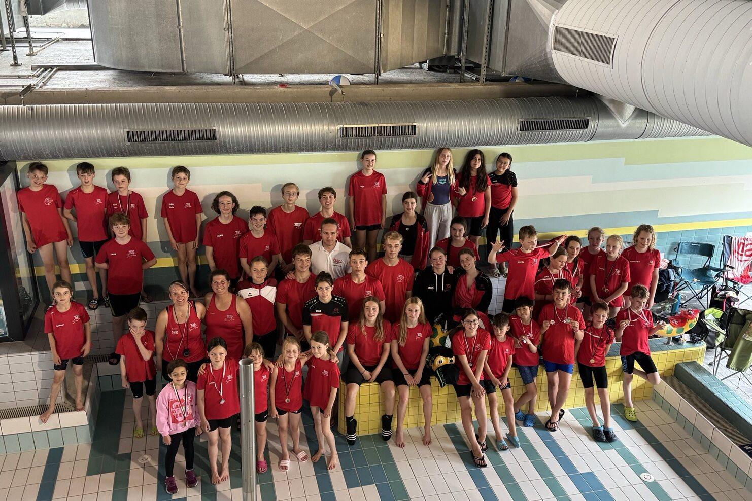 Gruppe von Schwimmern in roten T-Shirts posiert lächelnd am Beckenrand in einem Hallenbad.