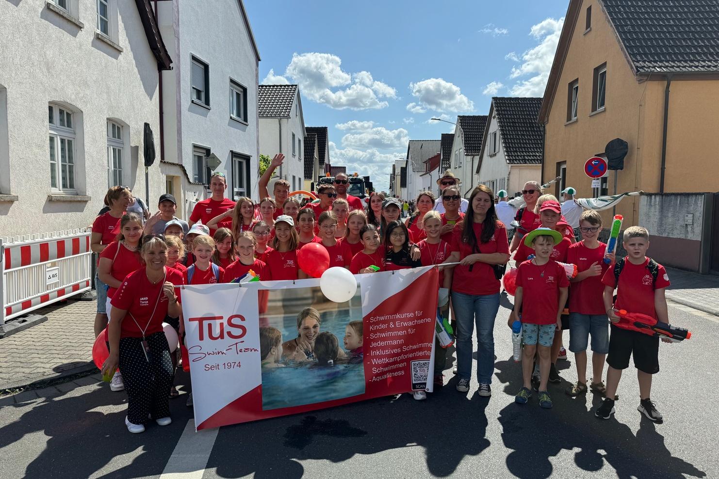 Gruppe von Kindern und Erwachsenen in roten T-Shirts mit Banner bei einem Umzug auf einer sonnigen Straße.