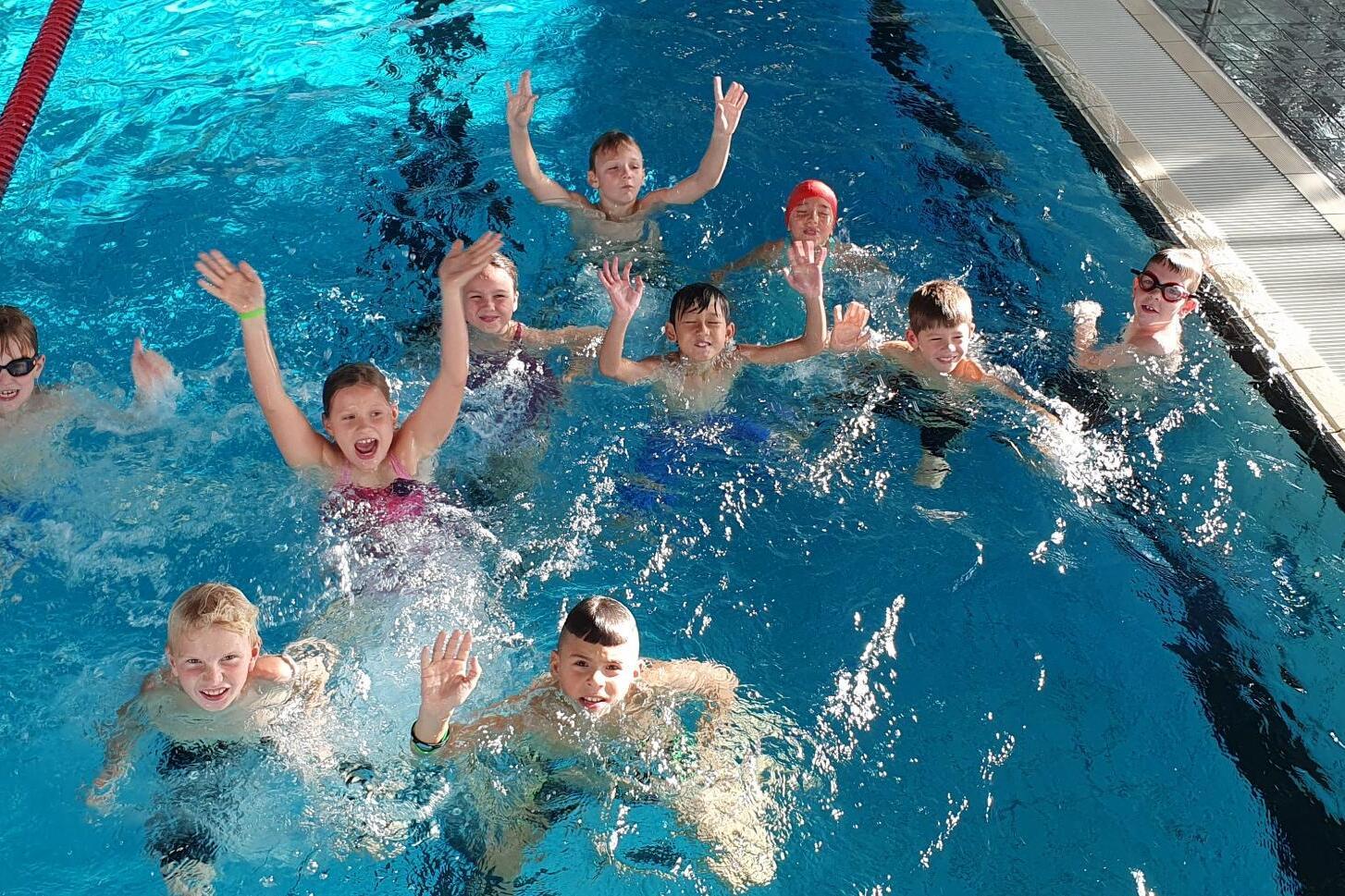 Gruppe von Kindern im Wasser, die fröhlich winken und Spaß im Schwimmbad haben.