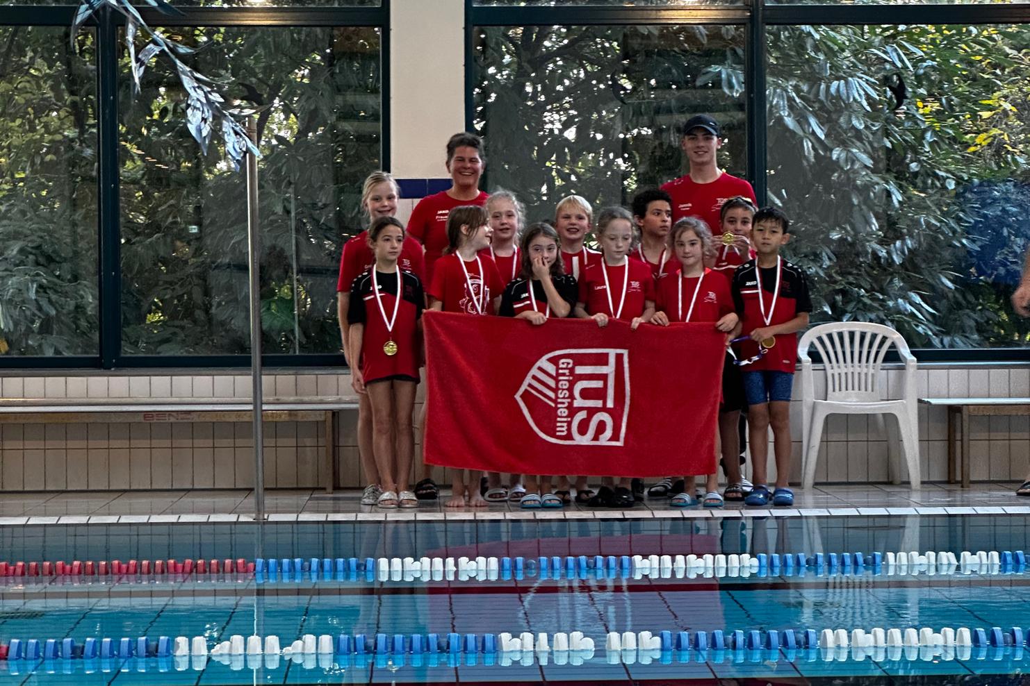 Gruppe junger Schwimmer in roten T-Shirts mit Medaillen, posiert vor einer Schwimmhalle mit einer Fahne.